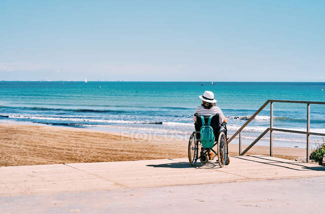 Повернення до нерозпізнаної жінки - мандрівника в інвалідному візку з рюкзаком, який насолоджується літньою подорожжю на пляжі біля блакитного моря. — стокове фото