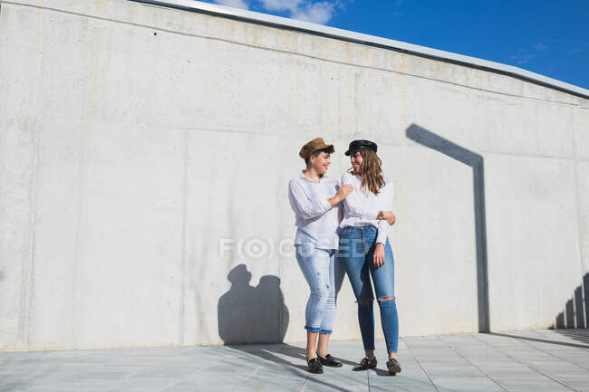 Full body of young positive female friends in trendy outfits and hats standing looking at each other on walkway near gray wall in sunny day under blue sky — Stock Photo