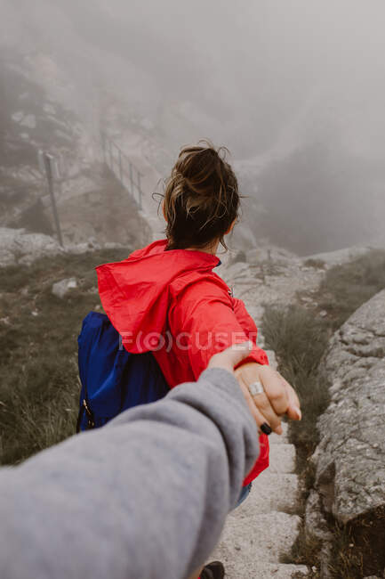 Back view of young tourist with backpack holding hand of human and walking on hill near footbridge — Stock Photo