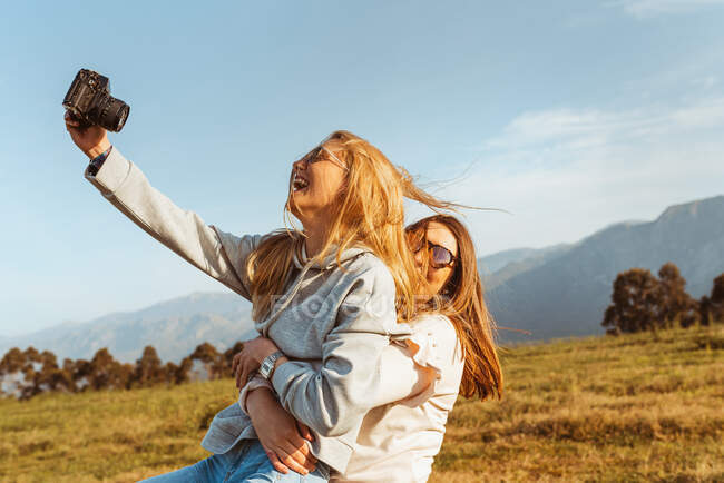 Cheerful woman carrying happy girlfriends in arms spending time together on field in high mountains in sunlight and taking selfie — Stock Photo