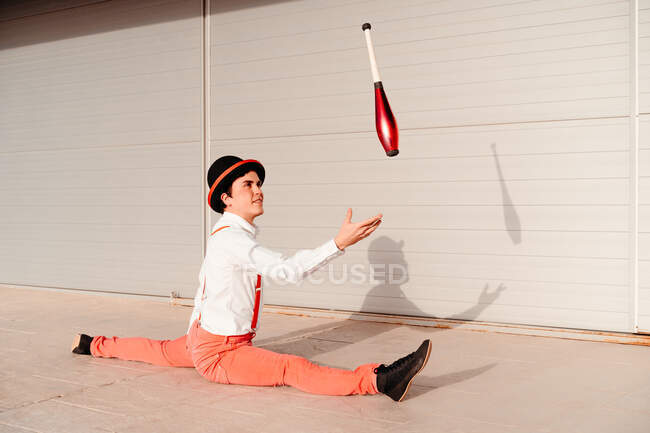 Ganzkörper-Seitenansicht eines talentierten flexiblen jungen männlichen Zirkusartisten, der Spagat macht und mit Schläger jongliert — Stockfoto