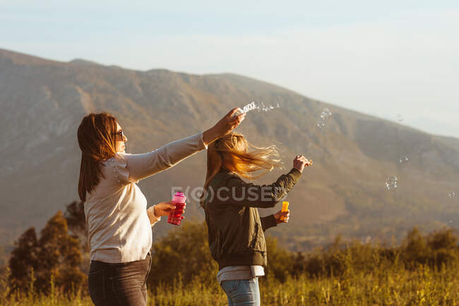 Fermer des amies dans des lunettes de soleil soufflant des bulles de savon ensemble debout sur la prairie dans les montagnes — Photo de stock
