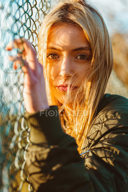 Mulher loira bonita em jaqueta olhando para a câmera de pé na cerca de elo de corrente em luz solar dourada — Fotografia de Stock