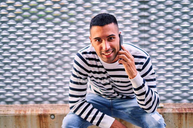 Froher hispanischer Mann in lässiger Kleidung, der lächelnd in die Kamera schaut und auf dem Smartphone spricht, während er an der Stadtmauer sitzt — Stockfoto