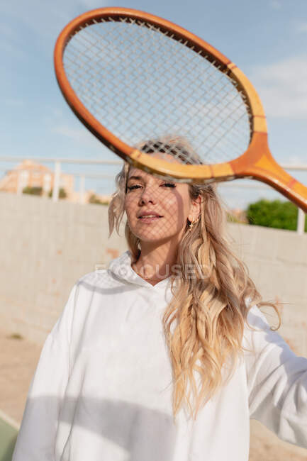 Позитивна молода жінка в білому одязі дивиться на камеру через тенісну ракетку, стоячи на сонячній вулиці — стокове фото