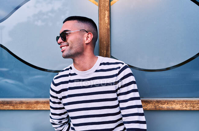 Позитивный взрослый латиноамериканец в полосатом свитере и стильных солнцезащитных очках улыбается и смотрит в сторону, опираясь на декоративную стеклянную стену на городской улице — стоковое фото