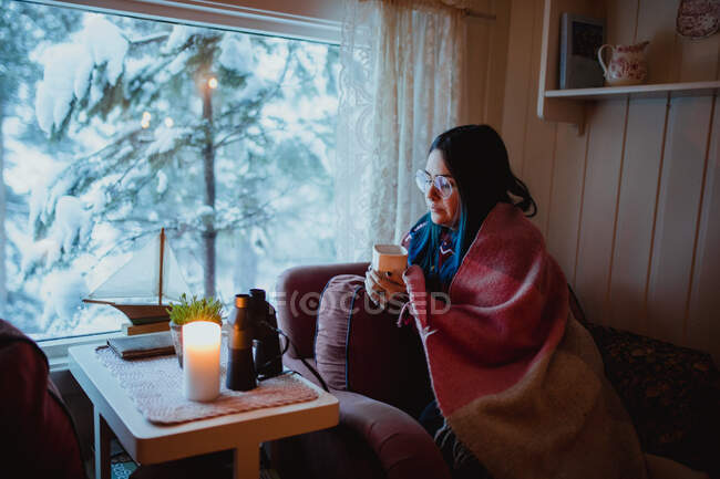 Vue latérale de la jeune femme en lunettes sous couverture reposant sur un canapé avec tasse près de la fenêtre avec vue sur les bois de sapin dans la neige — Photo de stock