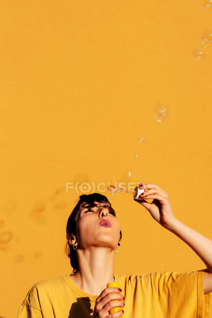 Femme moderne avec percing soufflant des bulles de savon sur une journée ensoleillée contre un mur jaune — Photo de stock