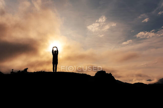 Silhouette of anonymous explorer with hands over head admiring mountainous terrain against cloudy sunrise sky in morning in nature — Stock Photo