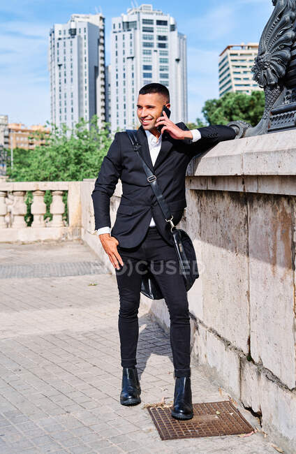 Full body Hispanic man in stylish outfit looking away and talking on cellphone while leaning on wall on city street — Stock Photo