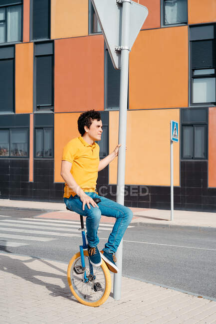 Вид сбоку на все тело молодого парня в повседневной одежде, сидящего на велосипеде на современной городской улице с разноцветным зданием — стоковое фото