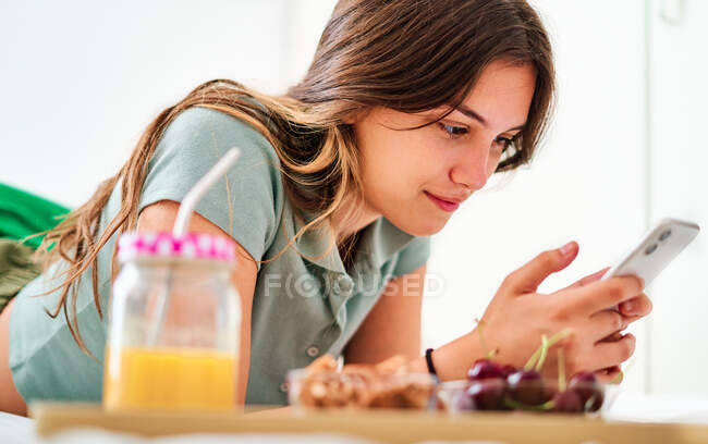 Vista lateral de una joven estudiante que navega por las redes sociales en el teléfono móvil cerca de la mesa con frutas frescas y jugo mientras pasa la mañana en casa - foto de stock