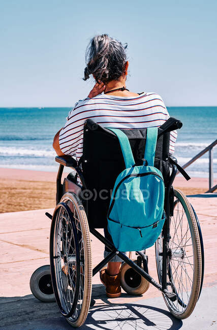 Back view of unrecognizable female traveler in wheelchair with backpack enjoying summer journey on beach near blue sea — Stock Photo