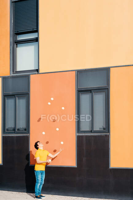 Corpo inteiro de homem jovem ágil em roupa casual realizando truque com bolas de malabarismo perto de edifício urbano moderno com paredes coloridas — Fotografia de Stock