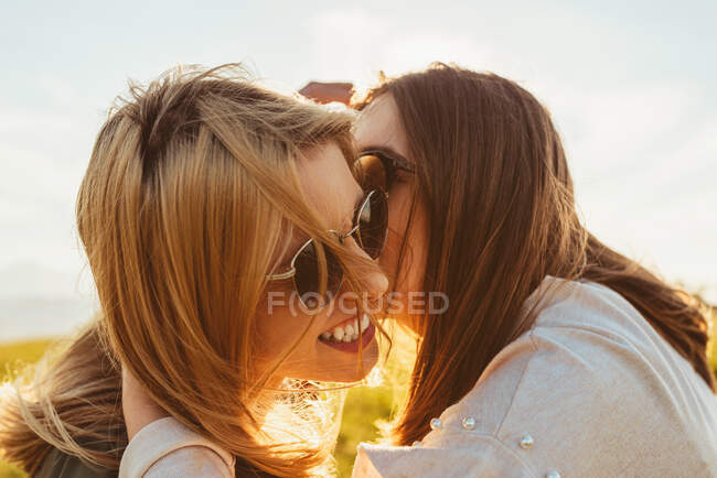 Счастливая брюнетка целует блондинку лучшую подругу в солнечных очках, сидящую в золотом солнечном свете на природе — стоковое фото