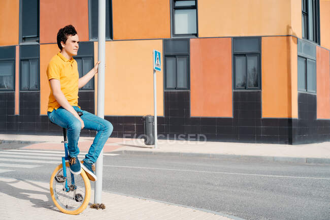 Full body side view of young guy in casual wear sitting on unicycle on modern urban street with colorful building — Stock Photo