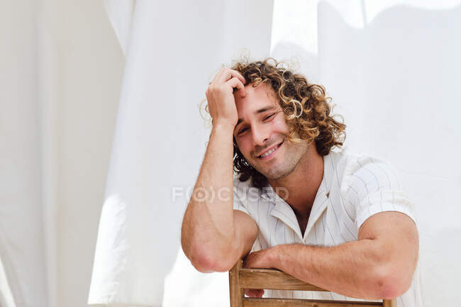Ravi beau mâle avec les cheveux bouclés assis sur la chaise et appuyé sur la main tout en regardant la caméra sur fond de rideaux blancs — Photo de stock