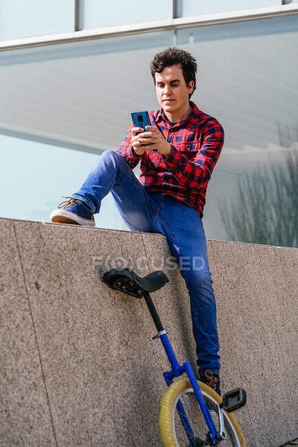 Повне тіло молодого хлопця в картатій сорочці і джинсах, що сидять на кам'яному паркані біля уніцикла і переглядають мобільний телефон у вільний час у місті — стокове фото