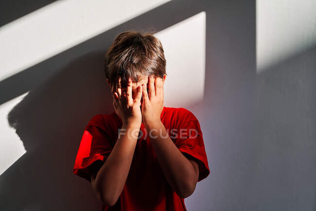 Расстроенный неузнаваемый мальчик, закрывающий лицо руками и плачущий, стоя у стены дома — стоковое фото