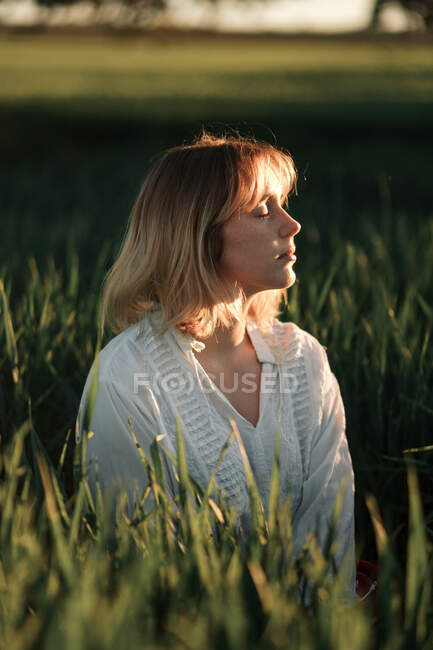 Mujer joven pacífica en blusa blanca de estilo retro sentada en medio de hierba verde alta y ojos cerrados mientras descansa en la noche de verano en el campo - foto de stock