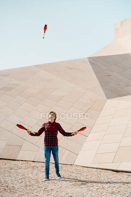 Full length of male performing trick with juggling clubs while standing against contemporary stone building with unusual geometric architecture — Stock Photo