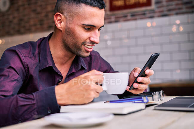 Счастливый латиноамериканец в фиолетовой рубашке наслаждается горячим напитком и просматривает сотовые сети во время перерыва во время работы в кафе — стоковое фото