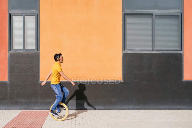Full body side view of modern young male in bright orange shirt and jeans listening to music through wireless headphones while riding unicycle on pavement near urban building — Stock Photo