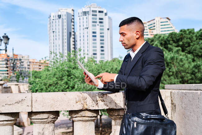 Vista lateral do jovem gerente executivo hispânico focado em terno elegante com informações de leitura de saco no tablet enquanto estava perto de trilhos no terraço urbano — Fotografia de Stock
