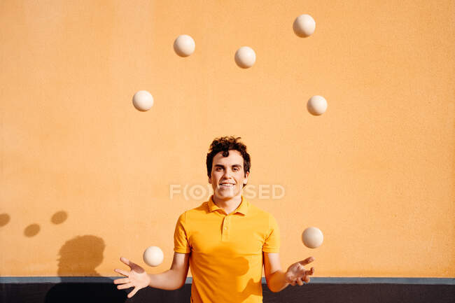 Feliz joven talentoso macho realizando truco con bolas malabares mientras está de pie mirando a la cámara en el pavimento cerca de la pared naranja brillante - foto de stock