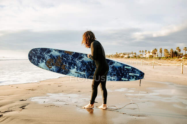 Side view of surfer man dressed in wetsuit walking with surfboard towards the water to catch a wave on the beach during sunrise — Stock Photo