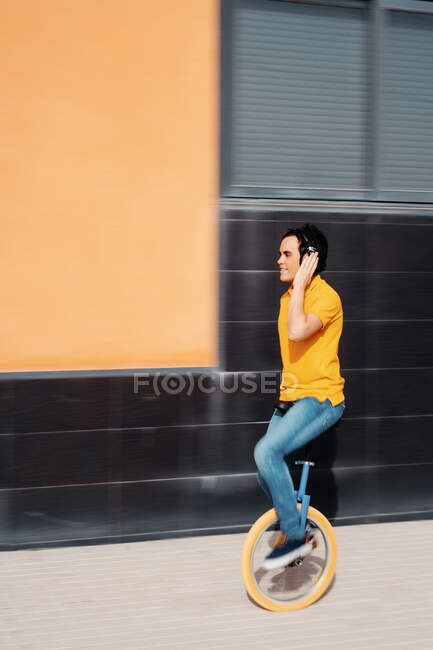 Vista lateral de cuerpo completo de un hombre joven moderno con camisa naranja brillante y jeans escuchando música a través de auriculares inalámbricos mientras monta monociclo en el pavimento cerca del edificio urbano - foto de stock
