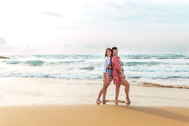 Full body of cheerful girlfriends in swimwear hugging each other while standing on sandy beach washed by waving sea — Stock Photo