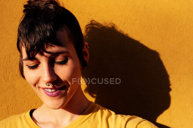 Femme moderne gaie avec une coupe de cheveux élégante et un sourire perçant avec les yeux fermés tout en se tenant près du mur jaune par une journée ensoleillée sur la rue — Photo de stock