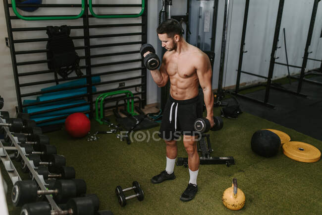 Treinador masculino jovem muscular com torso nu levantando halteres pesados durante o treinamento no ginásio — Fotografia de Stock