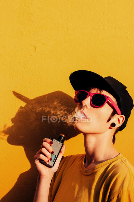 Mujer contemporánea en traje elegante exhalando humo mientras está de pie cerca de la pared amarilla y vapeando en la calle de la ciudad - foto de stock