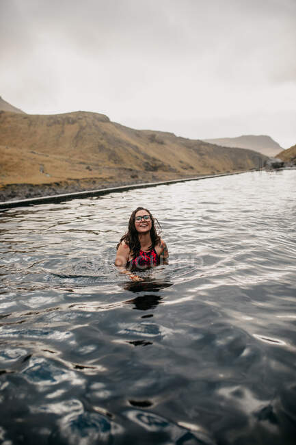 Young cheerful tattooed hipster in swimsuit in water between mountains — Stock Photo