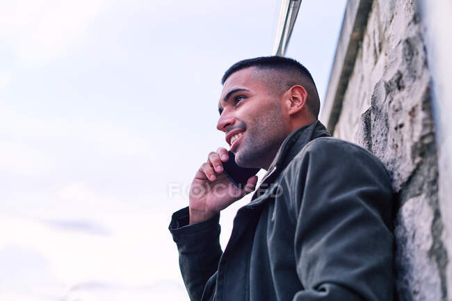 Baixo ângulo do homem hispânico feliz com café para ir sorrindo e olhando para longe, enquanto se inclina na parede de pedra e falando no celular na rua — Fotografia de Stock