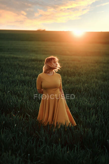Joven hembra en vestido vintage mirando hacia otro lado pensativamente mientras está de pie solo en el campo de hierba al atardecer en la noche de verano en el campo - foto de stock