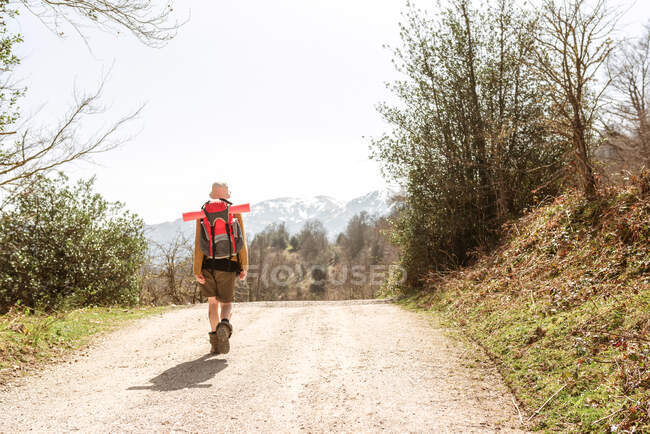 Vue arrière de l'explorateur avec sac à dos marchant sur une route sablonneuse menant vers les hauts plateaux par une journée ensoleillée — Photo de stock