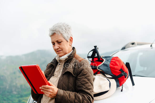 Elderly female traveler with short gray hair leaning on car and browsing tablet during road trip in countryside — Stock Photo