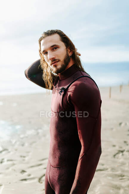 Vista laterale del giovane bel surfista uomo con i capelli lunghi vestito in muta in piedi guardando la fotocamera sulla spiaggia durante l'alba — Foto stock