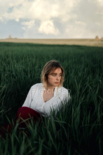 Jovem pacífica em retro blusa branca estilo sentado em meio a grama verde alta e olhando para longe enquanto descansa na noite de verão no campo — Fotografia de Stock