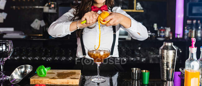 Cropped unrecognizable female barkeeper in stylish outfit decorating cocktail with lemon peel while standing at counter in modern bar — Stock Photo