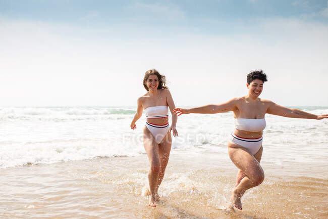 Allegre amiche in costume da bagno che corrono nell'oceano schiumoso vicino alla spiaggia sabbiosa sotto il cielo nuvoloso blu nella giornata di sole — Foto stock