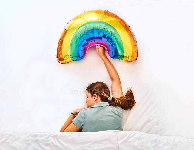 Вид зверху молода жінка тримає різнокольорову надувну повітряну кулю веселки під час сну під ковдрою в ліжку з білими простирадлами — стокове фото