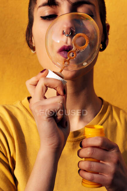 Mulher moderna com piercing soprando bolhas de sabão com olhos fechados na câmera no dia ensolarado contra a parede amarela — Fotografia de Stock
