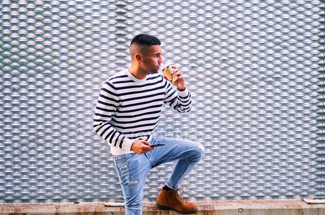 Adult Hispanic man in stylish casual clothes using cellphone and sipping coffee from takeaway cup while resting near ornamental gray wall on city street — Stock Photo
