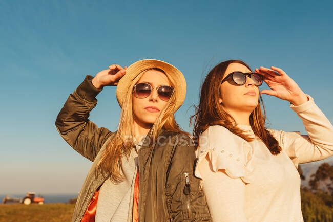 Jóvenes amigas íntimas con ropa elegante de pie juntas en el prado en las montañas mirando hacia otro lado en la luz dorada - foto de stock