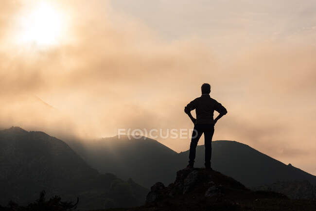 Silhueta de explorador anônimo com as mãos na cintura admirando terreno montanhoso contra céu nublado amanhecer na natureza — Fotografia de Stock