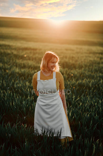 Giovane femmina in stile vintage grembiule bianco guardando via pensieroso mentre in piedi da solo in campo erboso al tramonto in estate sera in campagna — Foto stock